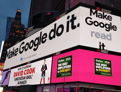 A bustling urban setting with large digital billboards displaying colorful advertisements. Prominent ads include 'Make Google do it' and 'American Idol Winner David Cook'. Bright neon colors and giant screens dominate the scene, hinting at a lively nightlife area.