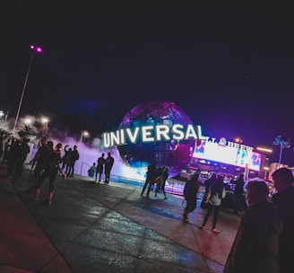 A large illuminated globe with the word 'Universal' is prominent at the center, surrounded by a misty, atmospheric effect. People are walking around the area, with some standing and looking at the globe. In the background, there are bright electronic billboards and palm trees, contributing to a lively and bustling environment.